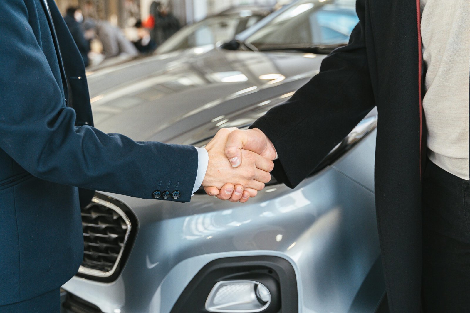 A close-up of two people shaking hands in front of a car in an indoor setting.